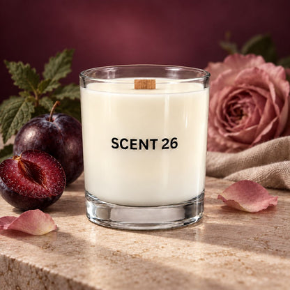 A clear glass candle jar filled with creamy off-white wax, labeled 'SCENT 26' in black text, set on a stone surface with plums, rose petals, and a pink rose in the background.