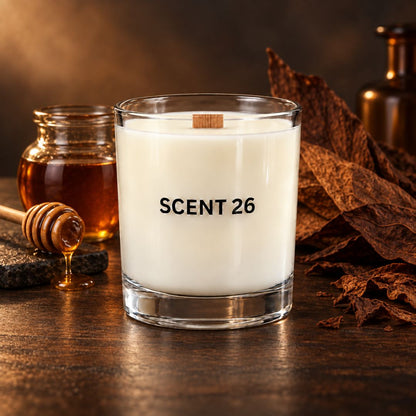 A clear glass candle jar labeled 'SCENT 26' filled with white wax, set on a dark wooden surface with a honey jar, honey dipper, and dried brown leaves around it.