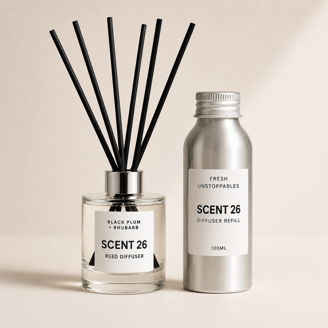 A reed diffuser set with a clear glass bottle, black reeds, and a silver metal diffuser refill bottle, both labeled SCENT 26, on a warm beige background.