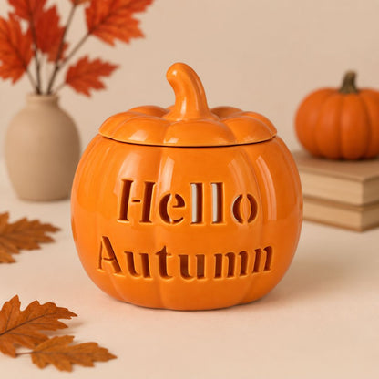 An orange pumpkin-shaped ceramic jar with a lid and the words 'Hello Autumn' embossed on the front, styled on a beige surface with fall leaves, a small vase, and a small pumpkin in the background.
